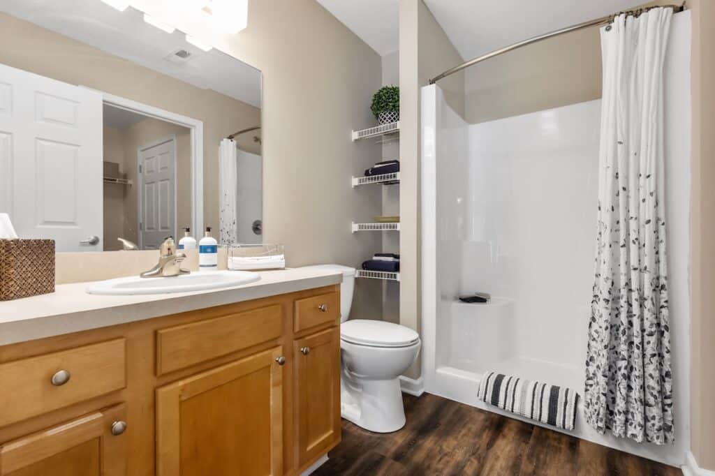 A bathroom with wood cabinets, a white sink, a toilet, open shelves with towels, and a shower with a white curtain and a striped rug on a wood-look floor.