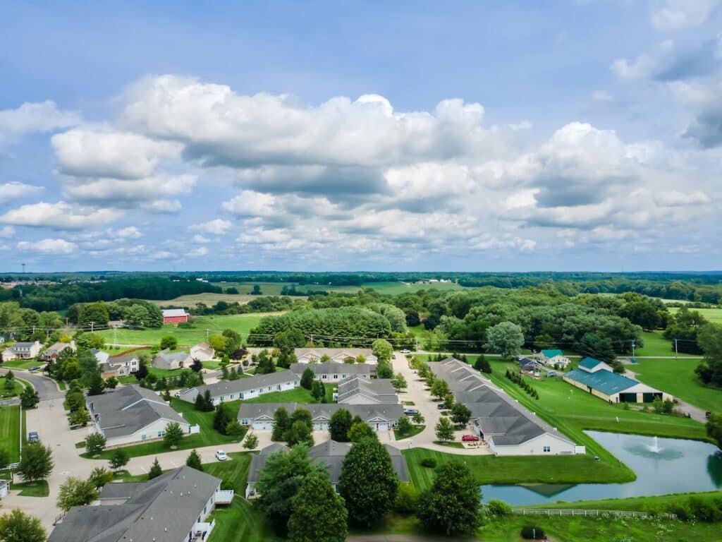 Aerial view of a suburban neighborhood with houses, trees, a pond, open fields, and a partly cloudy sky.