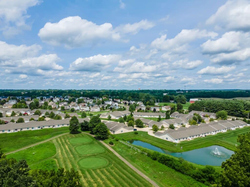 Aerial view of a suburban neighborhood with rows of houses, green lawns, a small pond with a fountain, and golf course features under a partly cloudy sky.