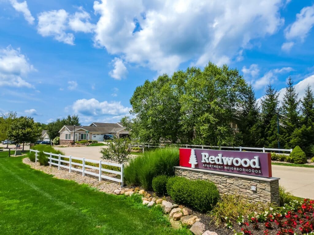 Entrance to Redwood Apartment Neighborhoods with a stone sign, landscaped greenery, a white fence, and residential buildings in the background under a partly cloudy sky.