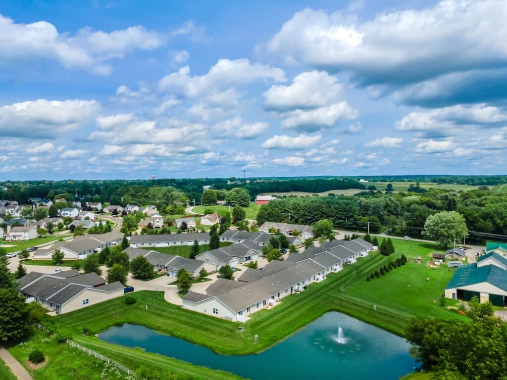 Aerial view of a suburban neighborhood with rows of houses, green lawns, a pond with a fountain, and open fields in the background under a partly cloudy sky.