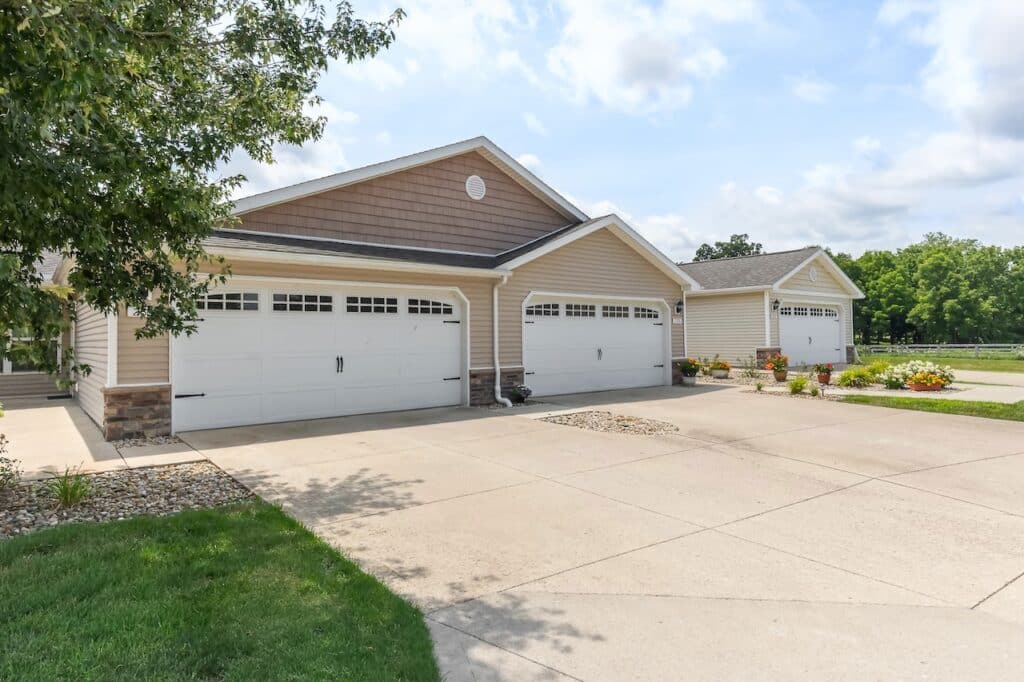 Three attached residential garages with white doors and beige siding are shown on a sunny day, with concrete driveways and small landscaped areas in front.