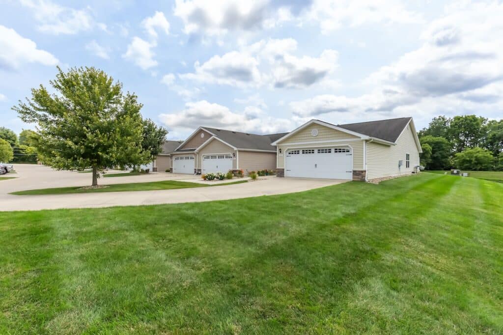 Single-story suburban homes with attached garages, well-maintained lawns, a large tree, and a curved driveway under a partly cloudy sky.