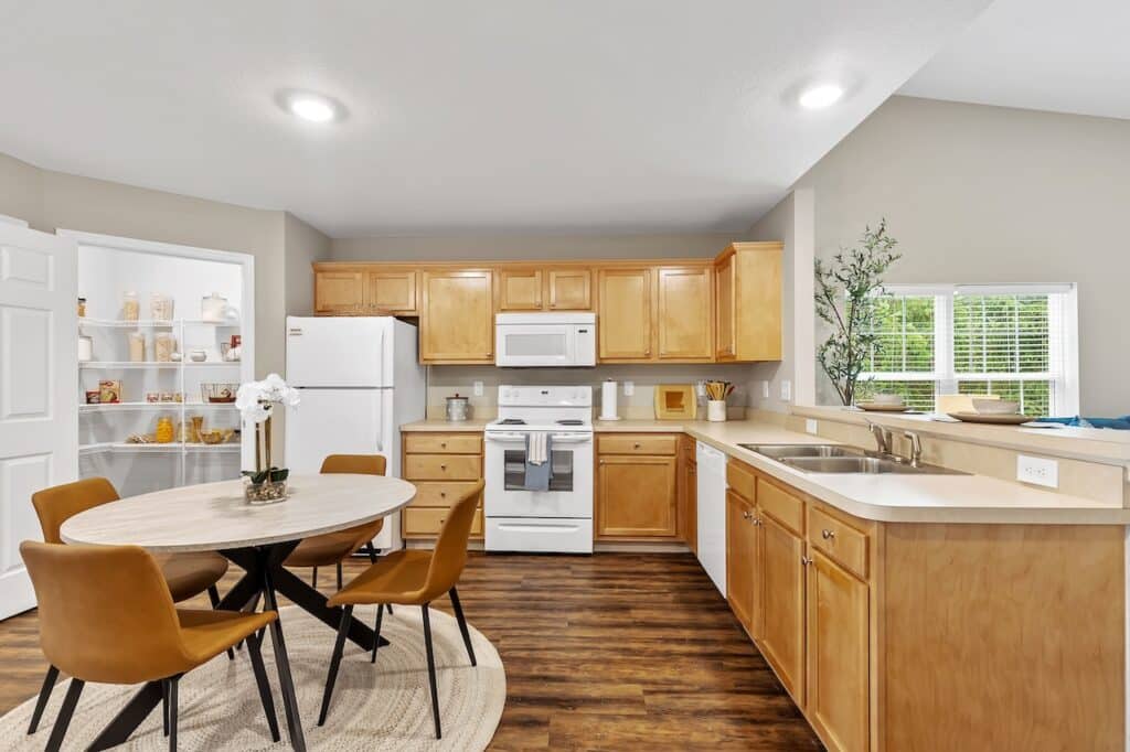 Modern kitchen with wooden cabinets, white appliances, and a round dining table with four brown chairs; pantry and window visible in the background.