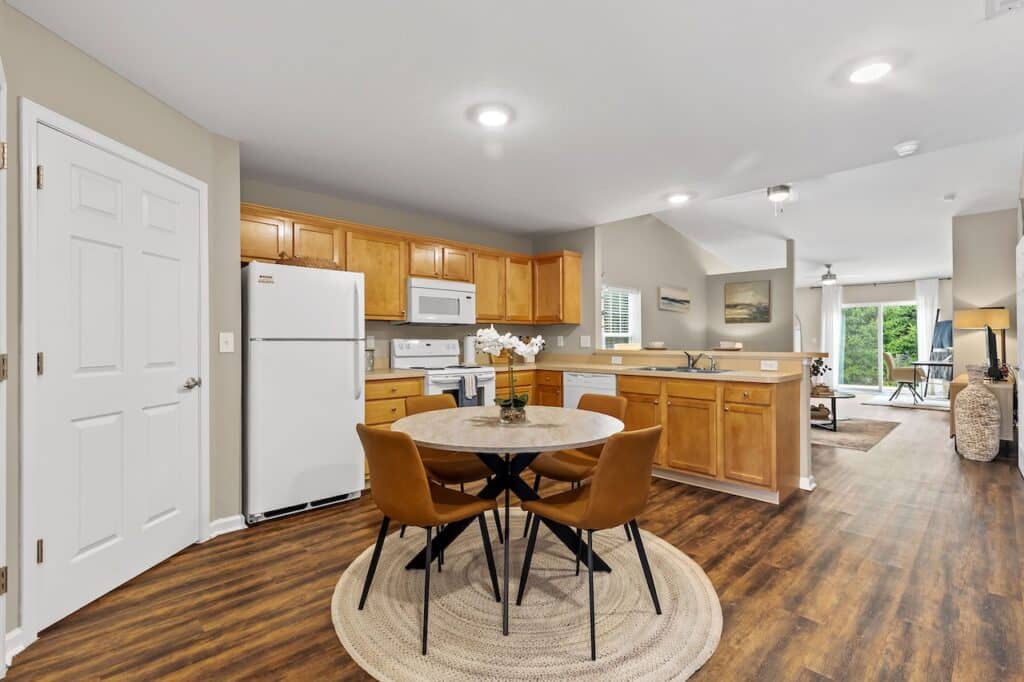 Open kitchen and dining area with wooden cabinets, white appliances, a round table with four chairs, and hardwood floors, leading to a living room and balcony.