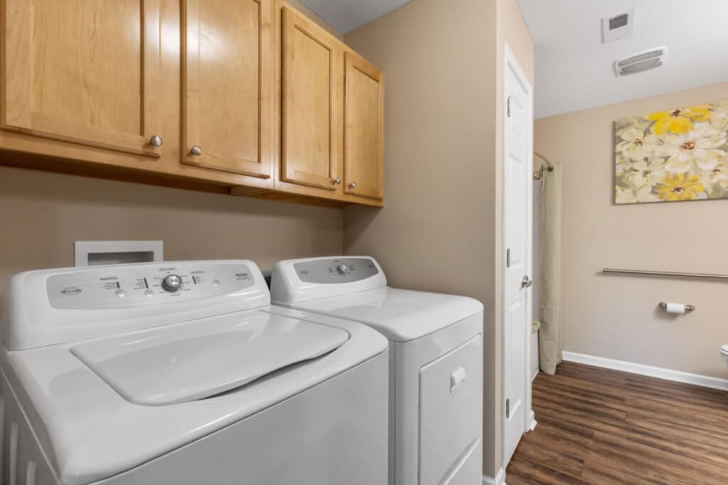 A laundry room with a washing machine and dryer, wooden cabinets above, beige walls, wood floor, and a bathroom visible in the background.
