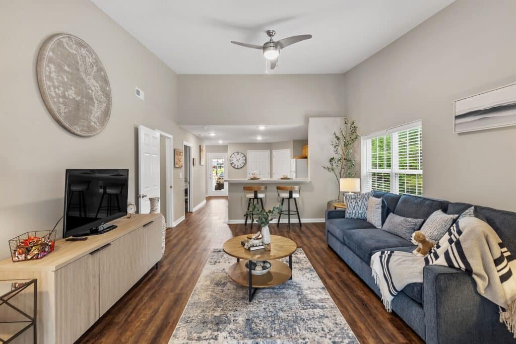 Modern living room with wooden floors, blue sofa, round coffee tables, TV on a light wood stand, and a kitchen with bar stools in the background. Bright window on the right.