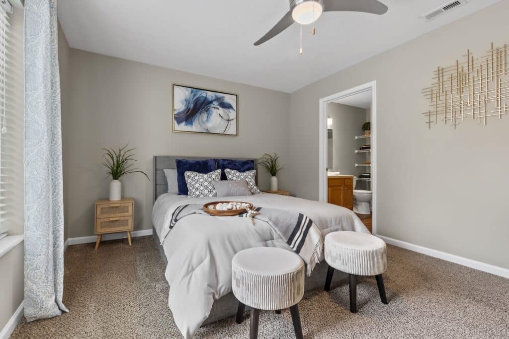 A neatly made bed with gray bedding and navy pillows sits in a bedroom with beige walls, two stools at the foot, and an open door revealing a bathroom.