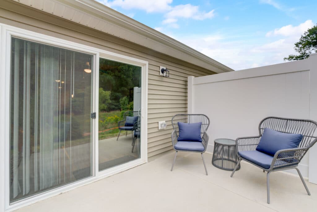 Patio area with two gray chairs with blue cushions and a small round table, next to sliding glass doors and a white privacy fence.
