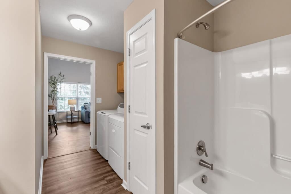 Laundry area with a washer and dryer next to a white bathroom bathtub; open door leads to a living space with a window and seating.