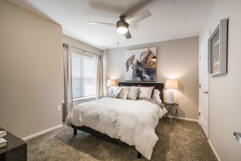 A bedroom with a neatly made bed, two bedside tables with lamps, abstract artwork above the headboard, a ceiling fan, and a window with light curtains.