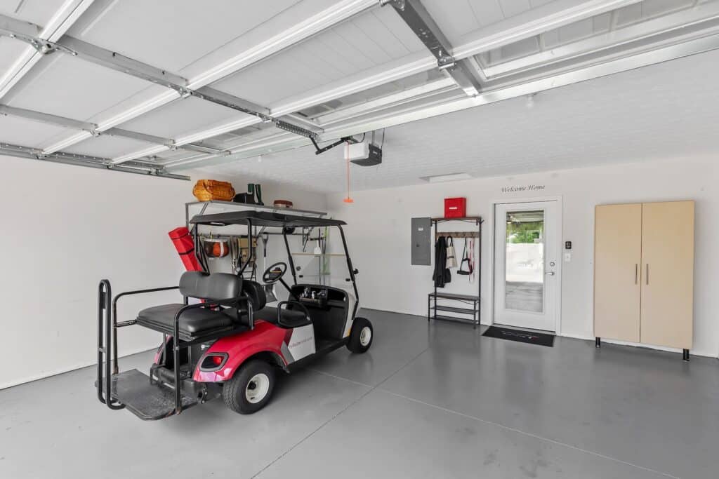 A clean, organized garage with a red and black golf cart parked inside, storage shelves and cabinets along the walls, and a white door leading outside.