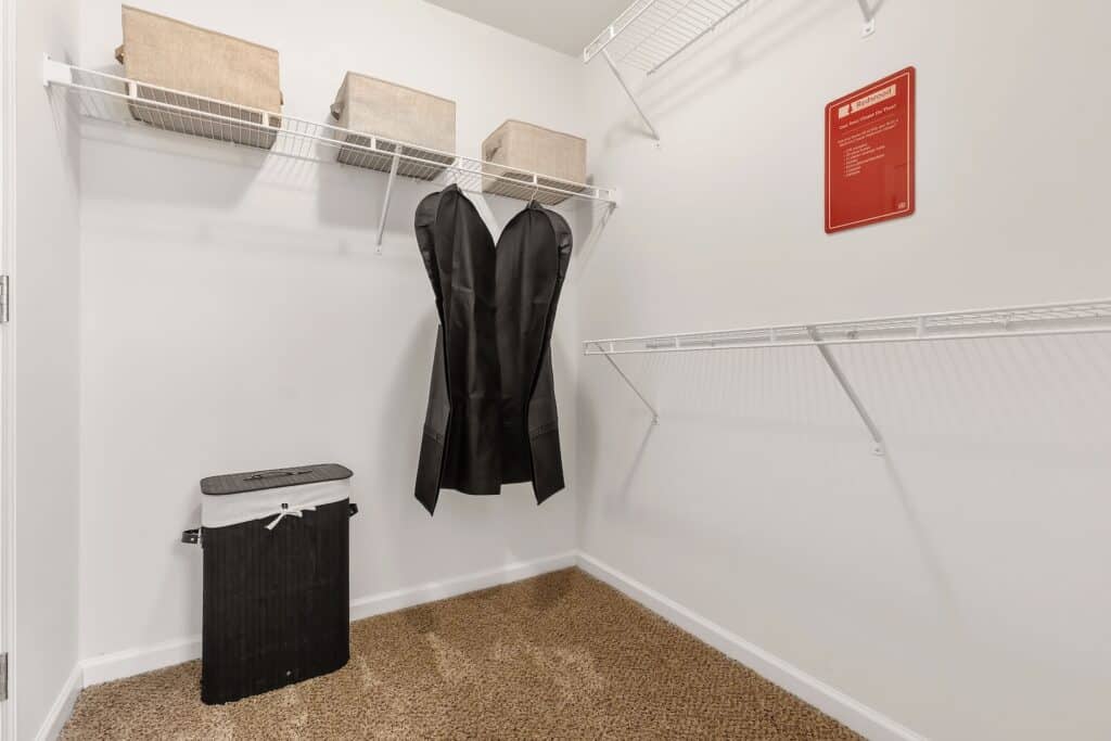 A walk-in closet with wire shelves, three fabric storage bins, a hanging garment bag, a black laundry hamper, and a red sign on the wall above beige carpet.