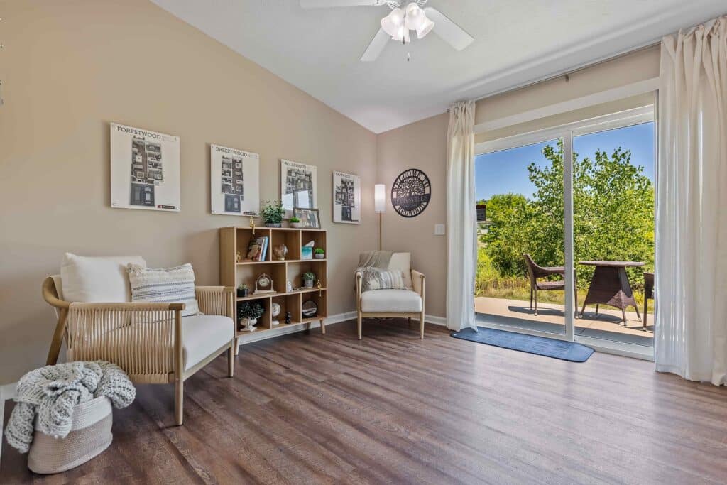 A bright living room with two chairs, a wooden bookshelf, framed wall art, and large glass doors opening to a patio with outdoor seating and greenery.