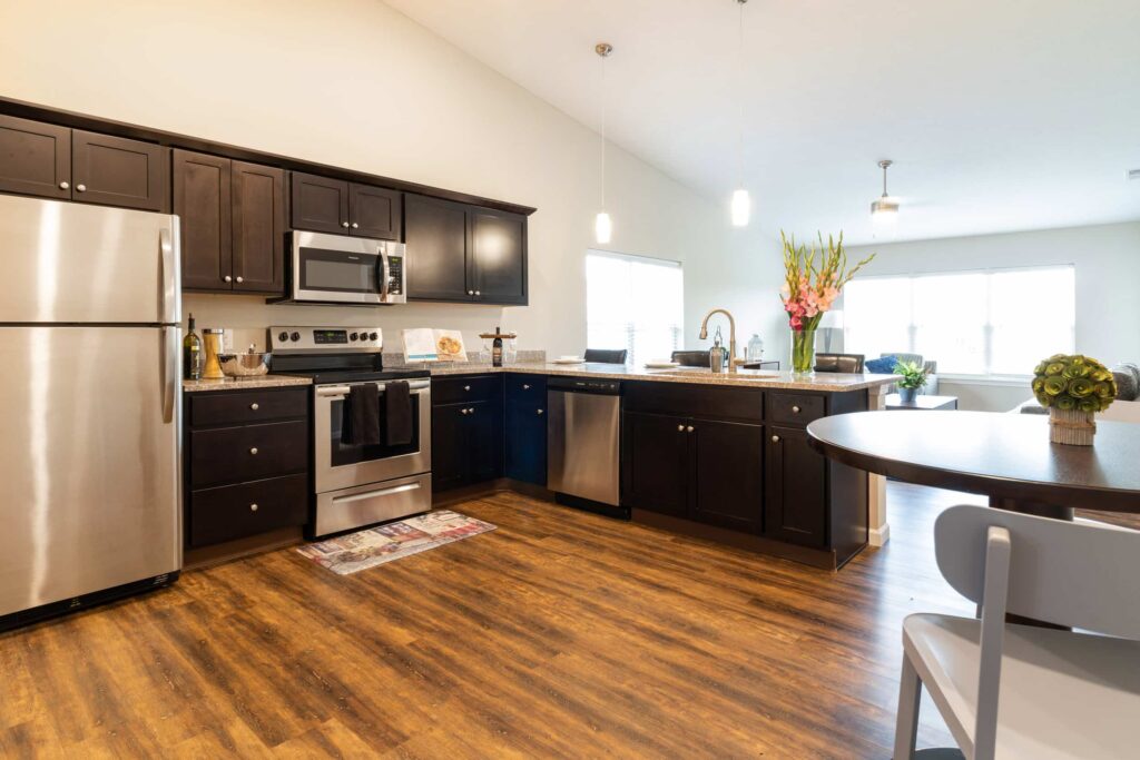Modern kitchen with dark cabinets, stainless steel appliances, a central island with flowers, and wood flooring. Dining table with chairs in the foreground. Bright, open space with natural light.