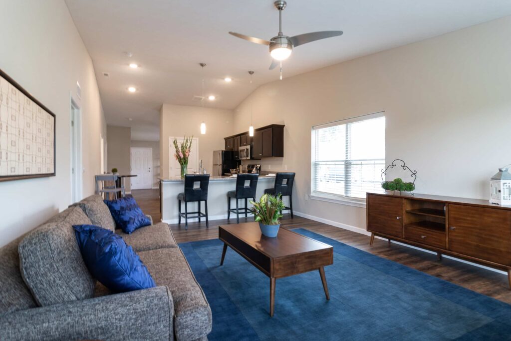 Modern living room with a gray sofa, blue cushions, wooden coffee table, blue rug, and open kitchen with barstools in the background. Ceiling fan and large window add light to the space.