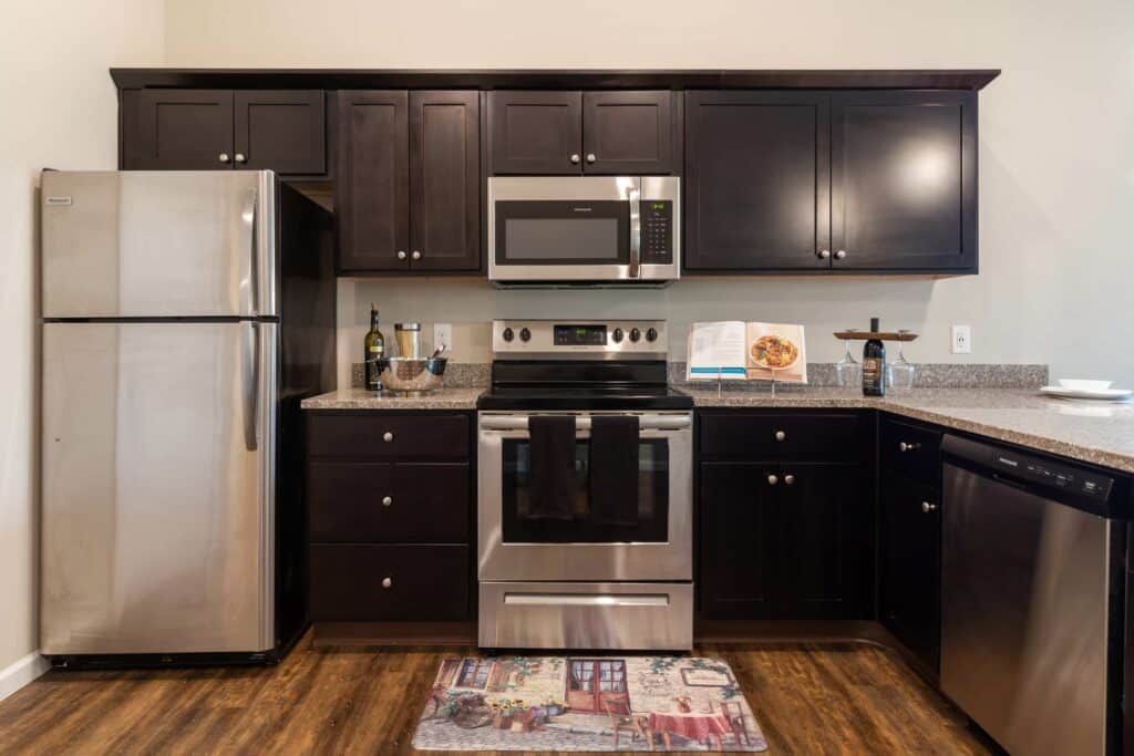 Modern kitchen with dark cabinets, stainless steel refrigerator, oven, microwave, and dishwasher; countertop holds a cookbook, bottles, and plates, with a colorful rug on the floor.