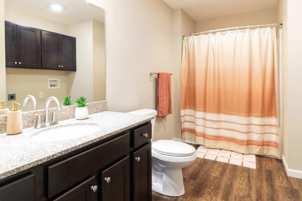 Modern bathroom with dark wood vanity, granite countertop, sink, toilet, orange towel, and orange ombre shower curtain on a curved rod; wood-patterned floor.