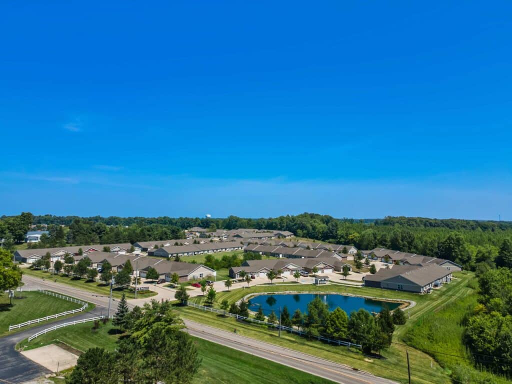 Aerial view of a residential neighborhood with rows of houses, a small pond, and surrounding greenery under a clear blue sky.