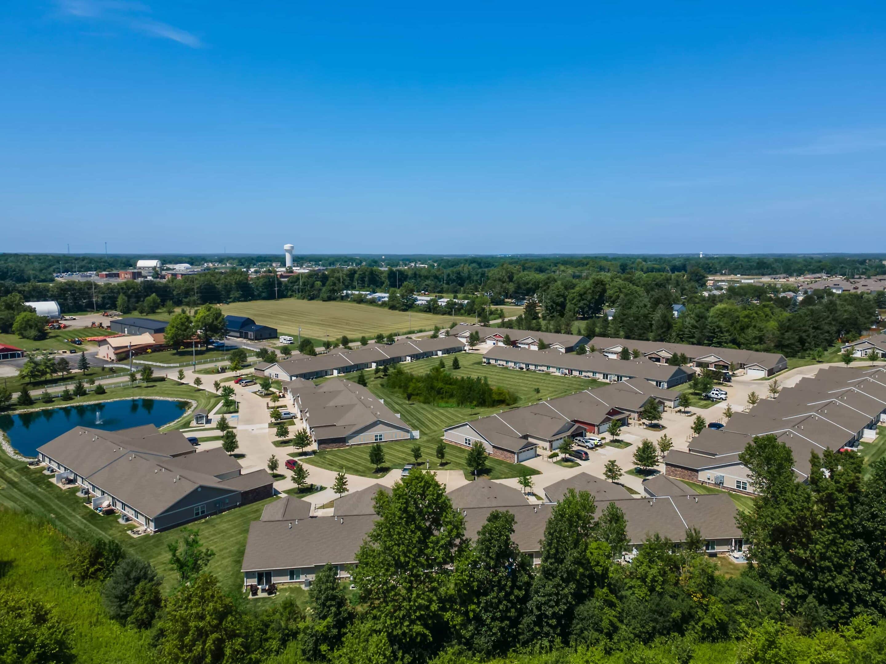 Aerial view of a suburban residential complex with multiple single-story buildings, a pond, surrounding greenery, and clear blue skies.