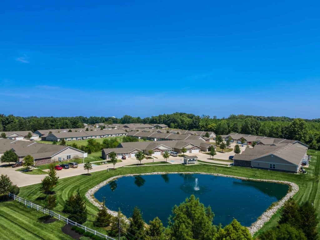 Aerial view of a residential community with single-story homes, manicured lawns, a pond with a fountain, and surrounding trees under a clear blue sky.