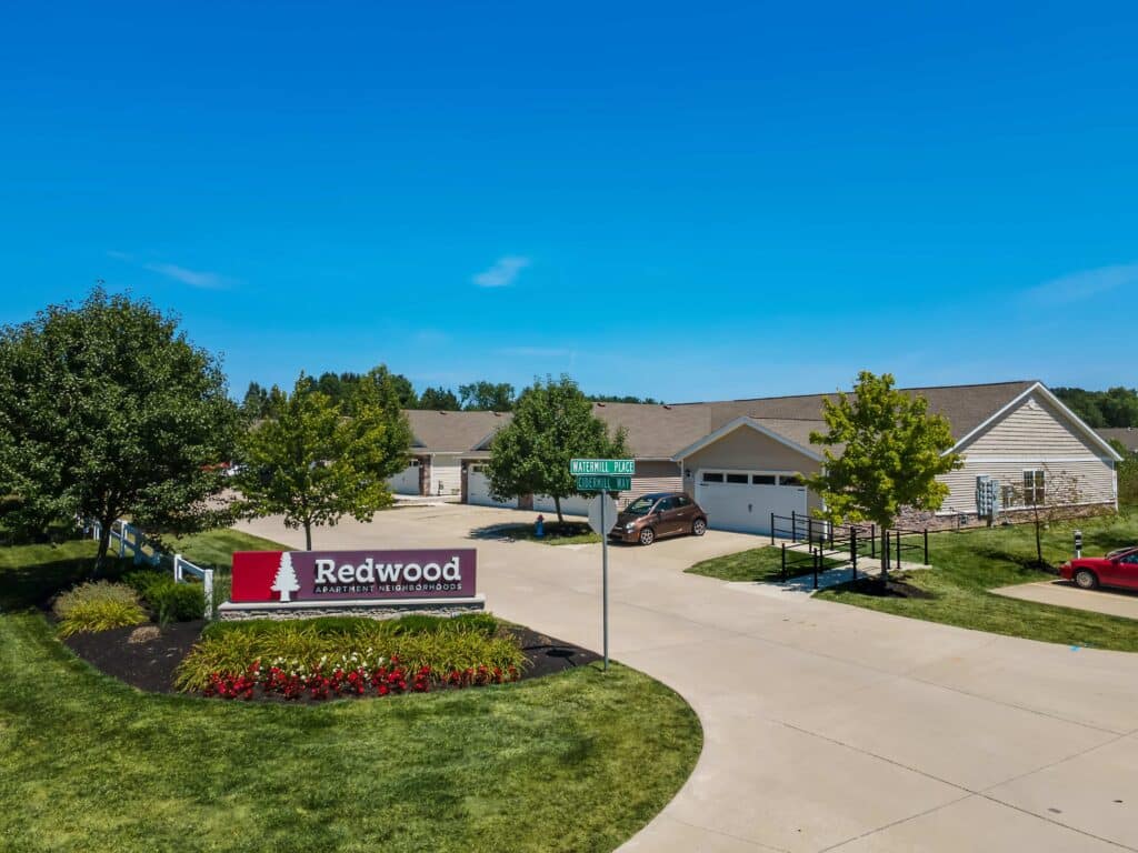 A residential complex with single-story homes, a driveway, parked cars, green lawns, and a “Redwood Apartment Neighborhoods” sign near a street intersection.