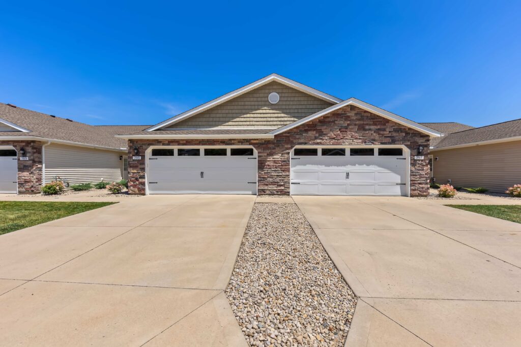 Two attached garages with white doors and stone facades, separated by a driveway with a central strip of rocks under a clear blue sky.