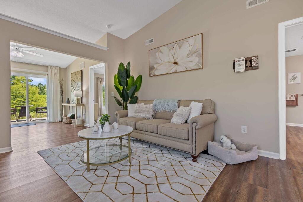 A living room with a beige sofa, white and gold coffee table, patterned rug, large plant, wall art, and a plush toy in a pet bed; dining area and outdoor view visible in background.