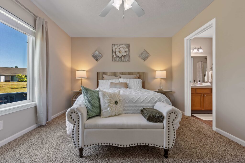 A neatly made bed with white bedding, two bedside tables with lamps, and a cushioned bench at the foot are in a carpeted bedroom with beige walls and an attached bathroom.