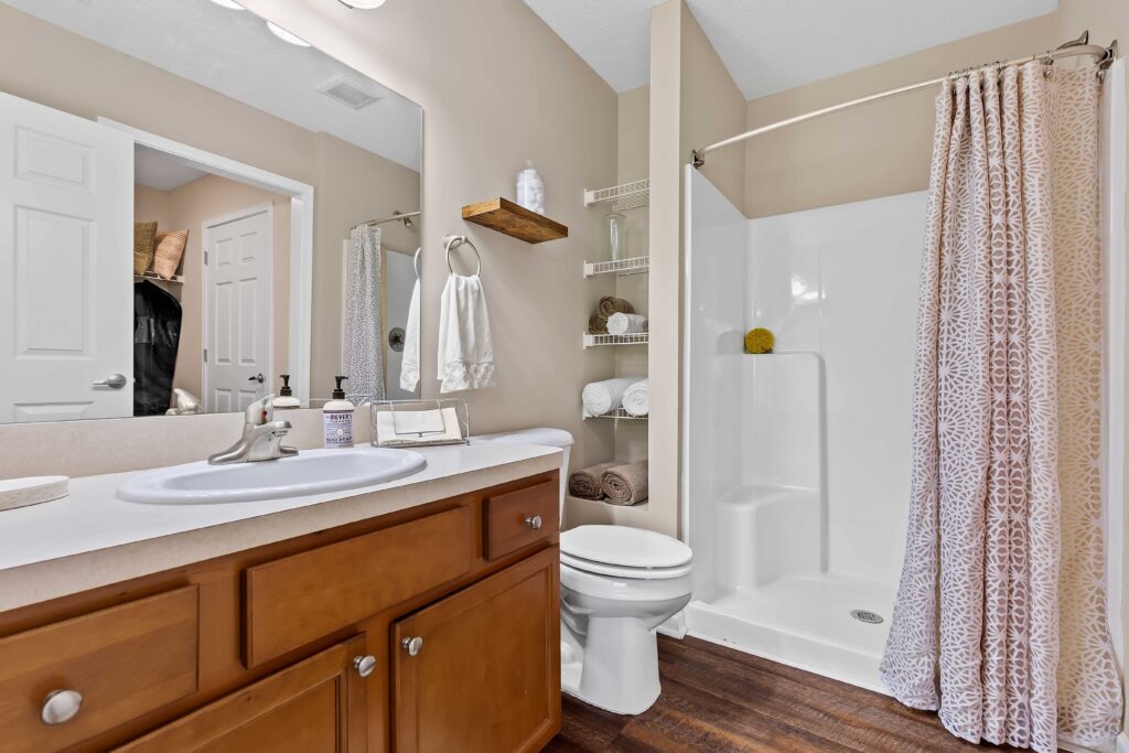 Bathroom with wood cabinets, single sink, white countertop, toilet, walk-in shower with patterned curtain, open shelving with towels, and neutral wall color.