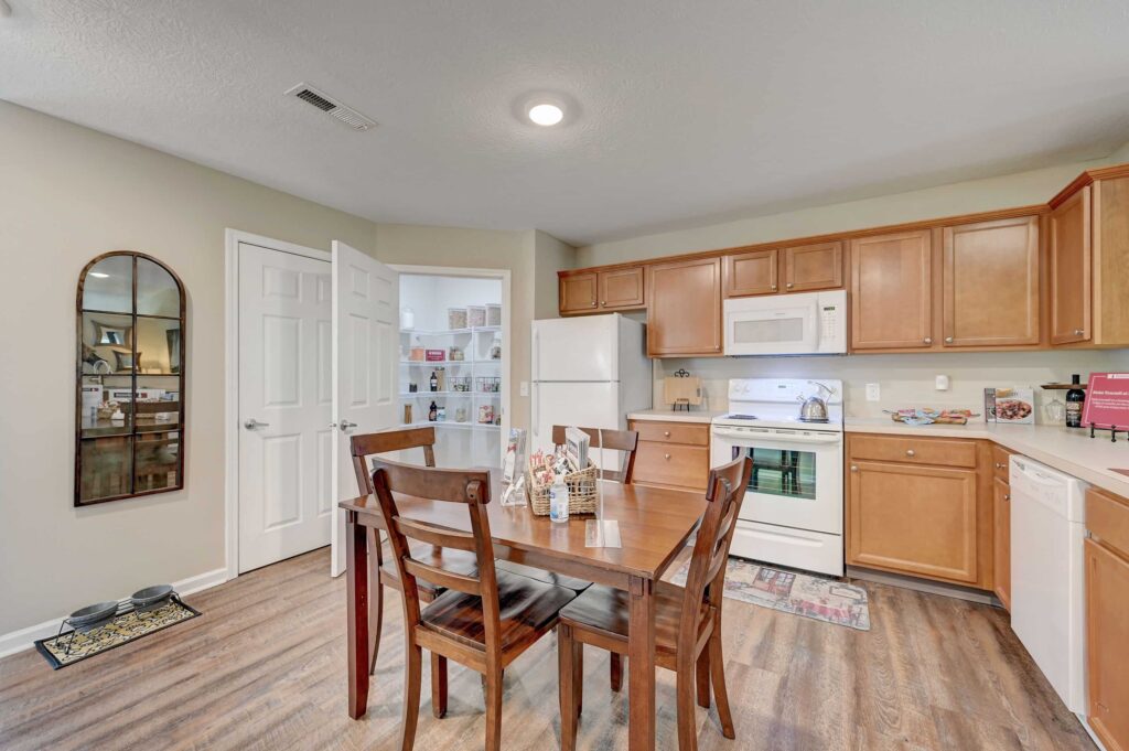 A kitchen with wooden cabinets, white appliances, a wooden dining table with chairs, and a pantry with an open door.
