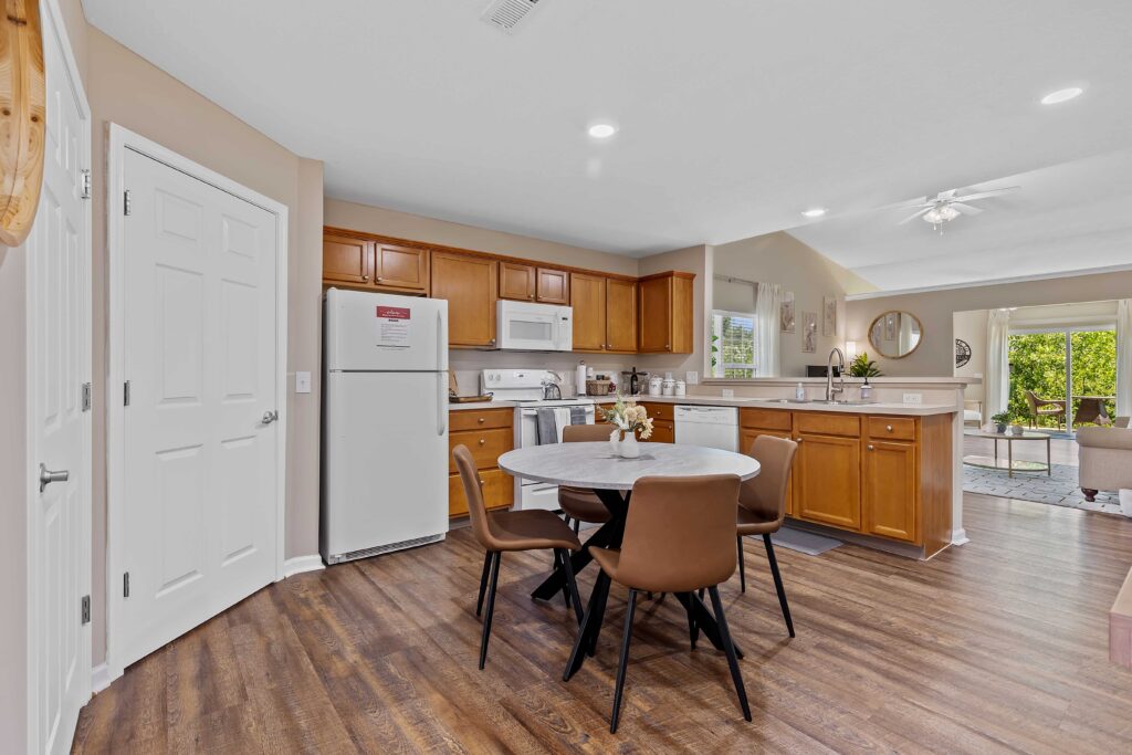 Modern kitchen with wood cabinets, white appliances, round dining table with four brown chairs, and an open layout leading to a living area with large windows.
