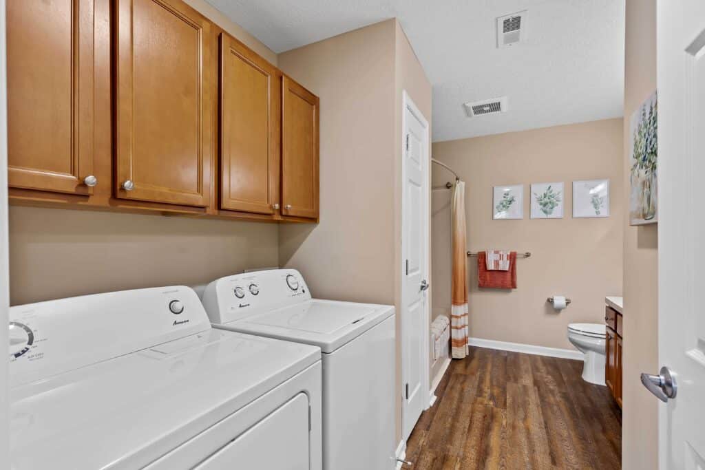 Laundry room with a washer and dryer, wooden cabinets above, and a view into a bathroom with a toilet, towel rack, and wall art. The floor is wood-style vinyl.
