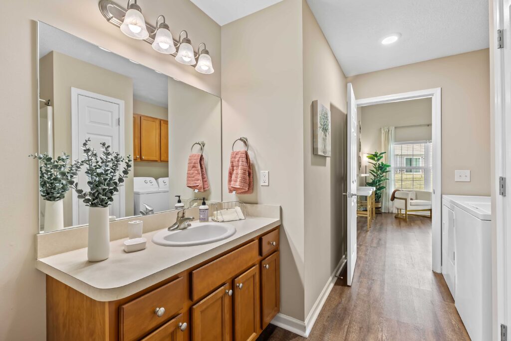 A bathroom with a vanity, sink, and mirror, featuring light wood cabinets, pink towels, and decorative plants, with a view into a laundry area and adjoining room.