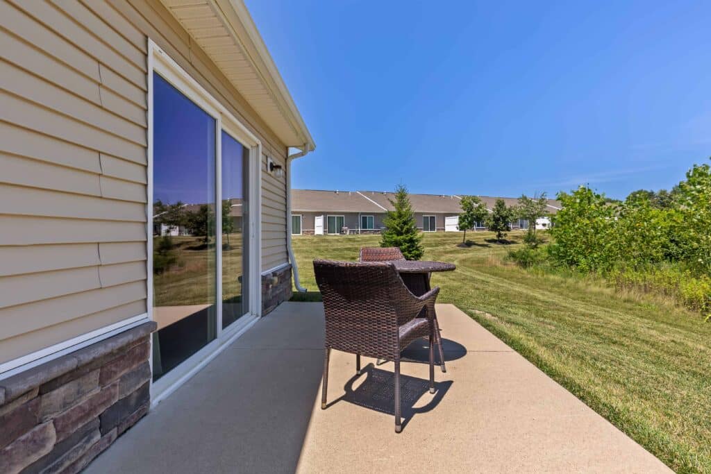 A small patio with a wicker table and two chairs is shown outside a house with tan siding, overlooking a grassy backyard and neighboring homes under a clear blue sky.