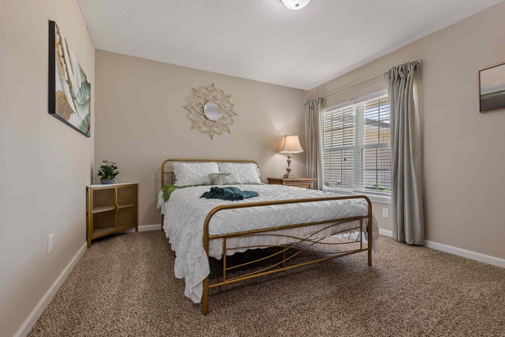 A neatly made bed with a metal frame is centered in a carpeted bedroom, with a nightstand, lamp, artwork, and a window with curtains letting in natural light.