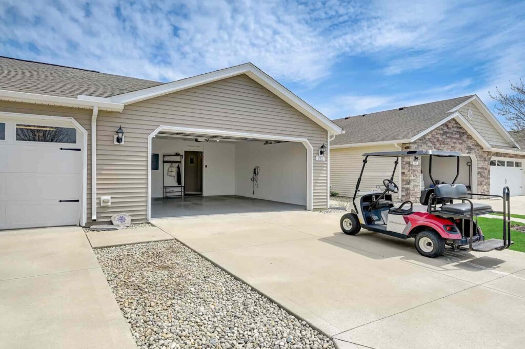 A beige house with an open two-car garage, a concrete driveway, and a red golf cart parked outside. The garage interior is empty and clean.