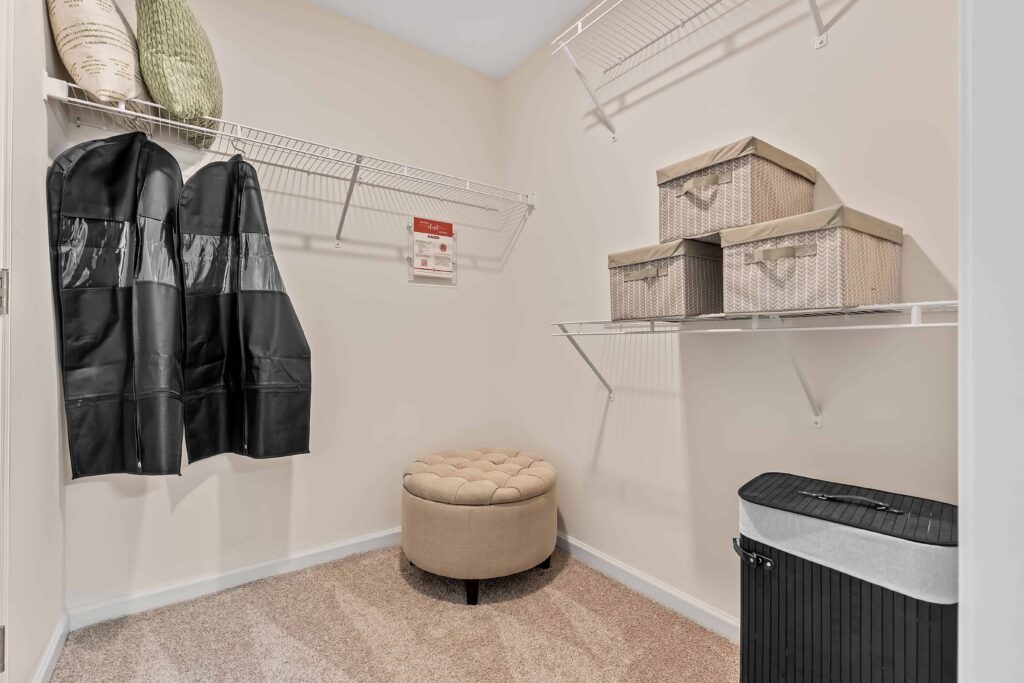 A walk-in closet with two black coats, woven baskets on shelves, a round beige ottoman, and a black laundry hamper on carpeted floor.