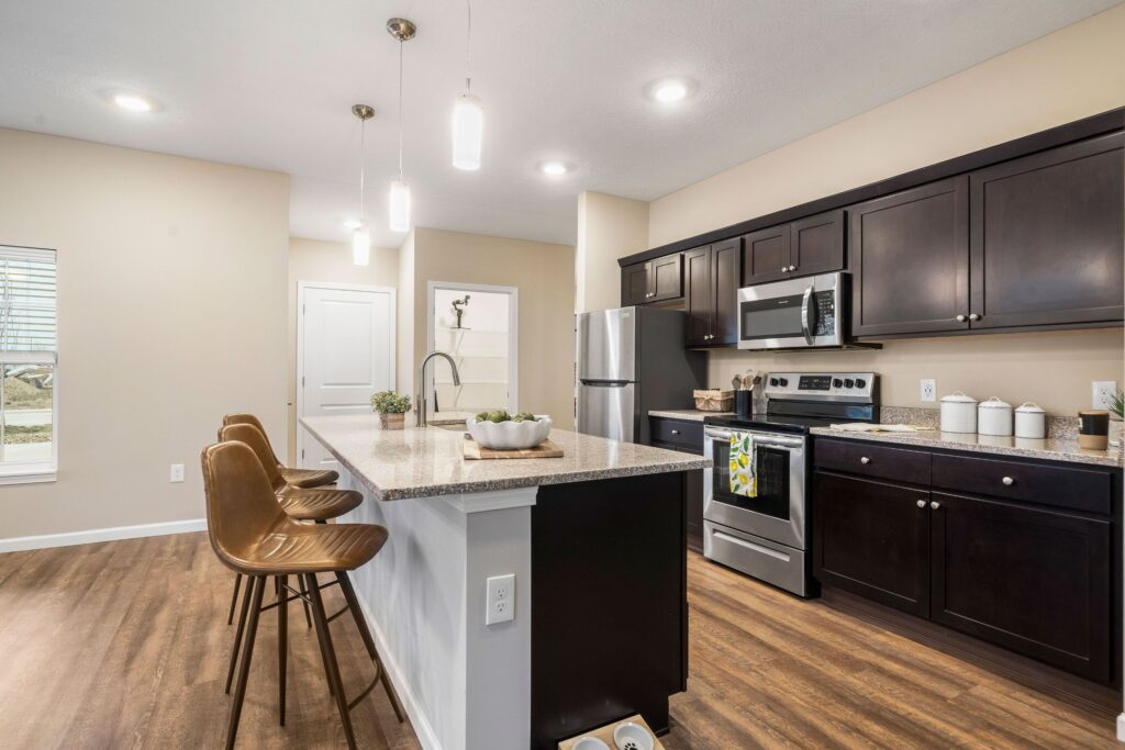 Modern kitchen with dark cabinets, stainless steel appliances, an island with three brown stools, and wood flooring under recessed and pendant lighting.