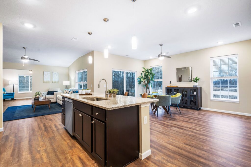 Open-concept living space with a kitchen island, dining area, and seating area. Wood floors, large windows, and modern lighting are visible throughout the room.
