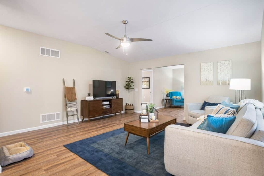 A modern living room with a light gray sofa, blue accents, a wooden coffee table, TV on a console, and hardwood floors. A ceiling fan and floor lamp are also visible.