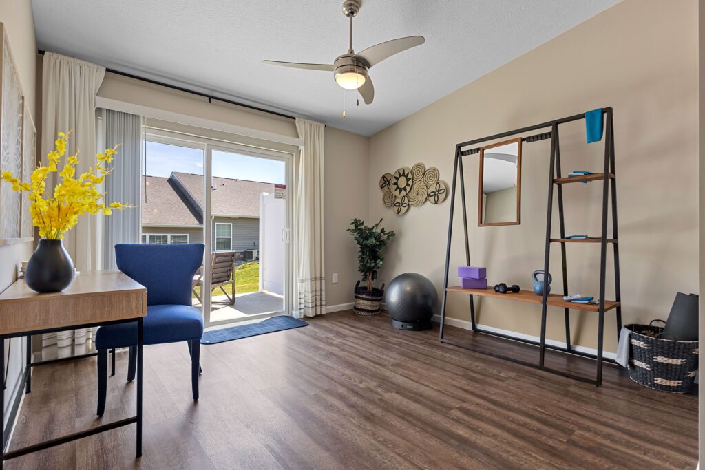 A bright room with wood flooring, a desk and chair, exercise equipment, shelves with decor, and sliding glass doors leading outside.