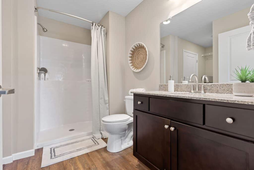 Modern bathroom with a walk-in shower, white shower curtain, toilet, dark wood vanity, dual sinks, granite countertop, and decorative accents on a wood floor.
