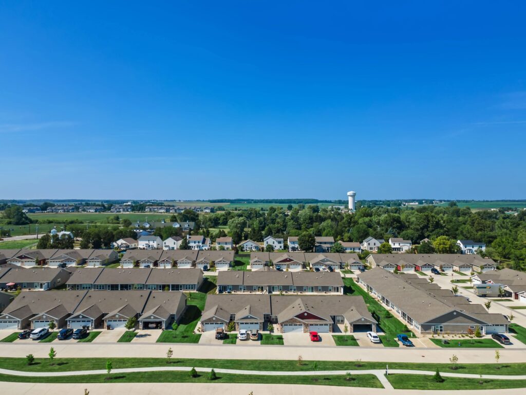 Aerial view of a suburban residential neighborhood with rows of houses, green lawns, a water tower in the distance, and open fields under a clear blue sky.