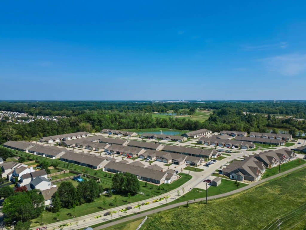 Aerial view of a suburban residential neighborhood with rows of townhouses, green spaces, roads, and surrounding trees under a clear blue sky.