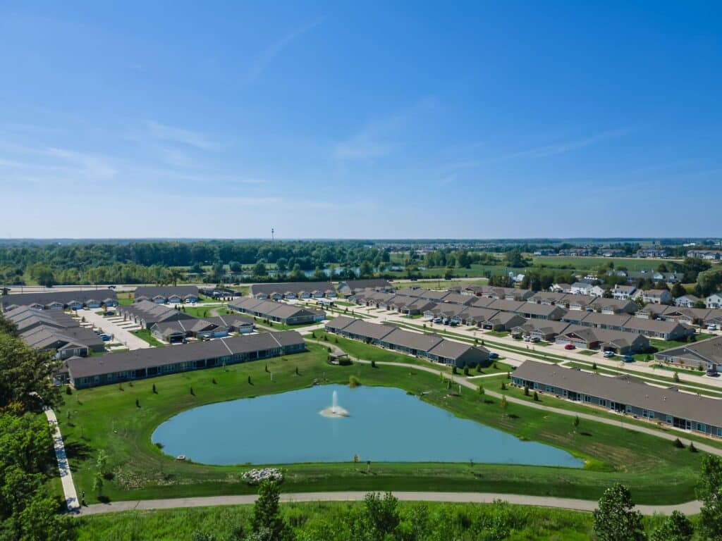 Aerial view of a suburban neighborhood with rows of houses, a small pond with a fountain, and green lawns under a clear blue sky.