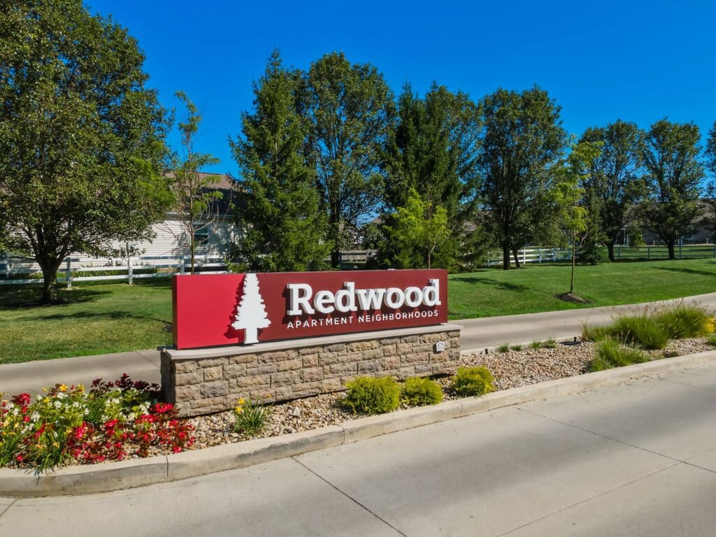 A red and white sign reading "Redwood Apartment Neighborhoods" is displayed in front of landscaping and a driveway, with trees and a white fence in the background.