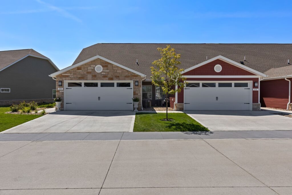 Two neighboring single-story houses with attached two-car garages and a small tree between their driveways under a clear blue sky.