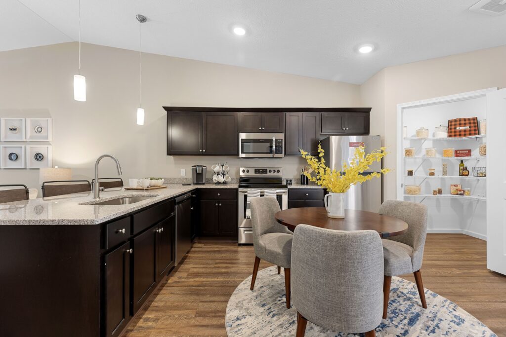 Modern kitchen with dark cabinets, stainless steel appliances, a round dining table with four chairs, and an open pantry stocked with food items in the background.