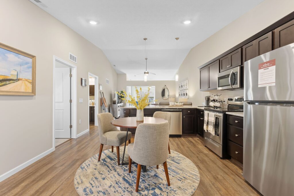 A modern kitchen and dining area with dark cabinets, stainless steel appliances, a round table with four chairs on a rug, and decorative accents throughout the room.